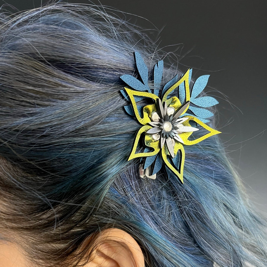An iridescent gold flower barrette with blue leaves, shown on a woman with blue hair