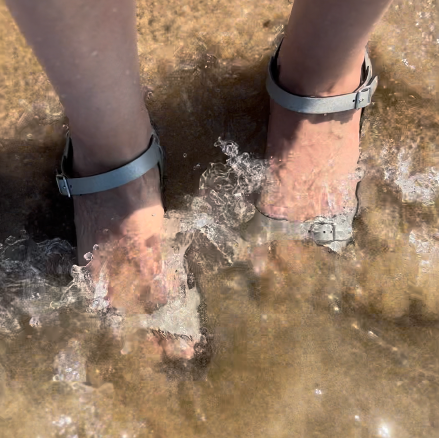 Ankle strap sandals in champagne color, shown on a model standing in the water as waves rush over her feet