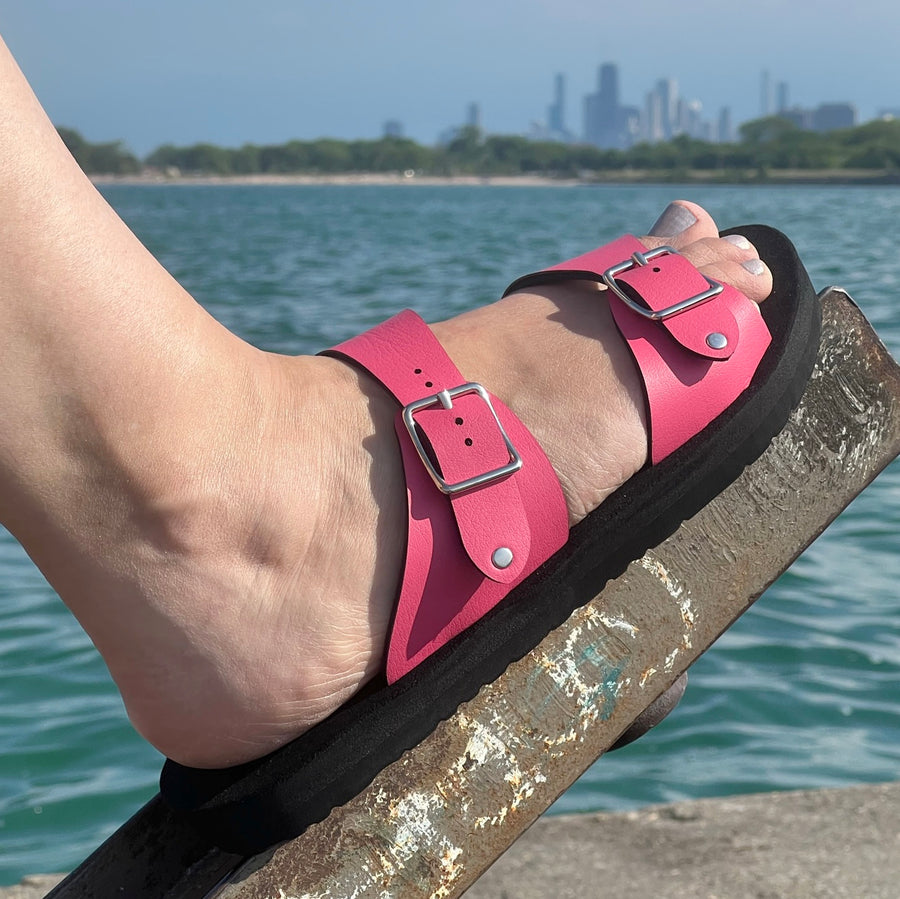 Side view of a flat, padded vegan sandal in hot pink, shown on a model with Lake Michigan and the Chicago skyline in the background