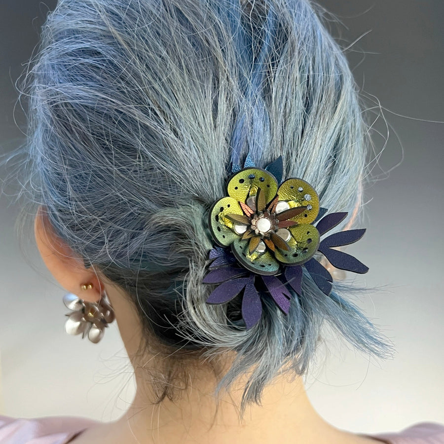 A thick elastic hair tie featuring a large iridescent gold faux leather flower accented by iridescent blue leaves shown on a model
