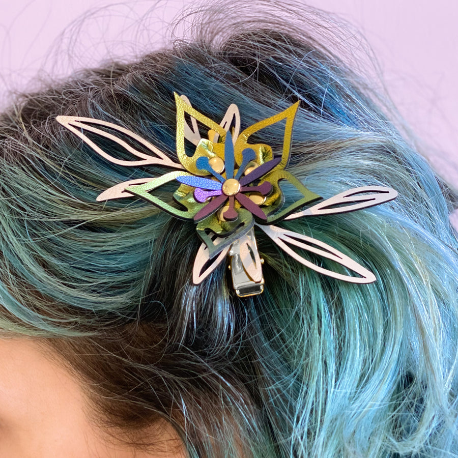A vegan leather barrette in clipped in turquoise hair, featuring large silver leaves and an iridescent gold spikey flower.