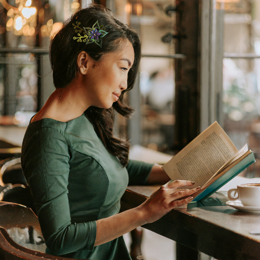 An iridescent green flower barrette with green-gold iridescent loopy leaves, shown on a woman in a green dress reading a book in a cafe