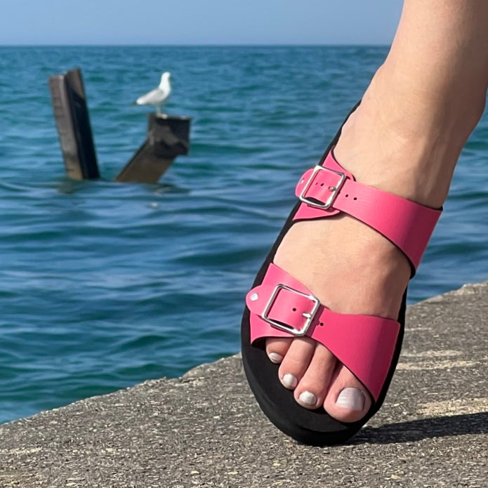 Side view of a flat, padded vegan sandal in hot pink, sown on a model with a backdrop of a seagull and Lake Michigan