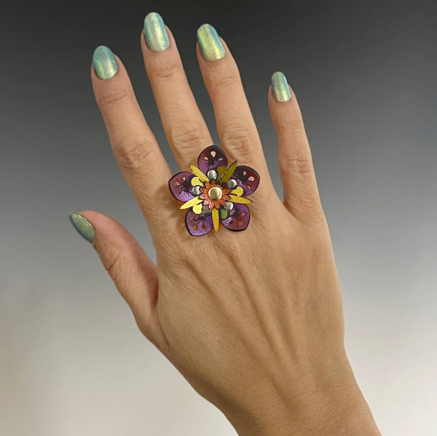 A faux leather ring with a purple iridescent flower and gold iridescent stamen on a model's hand
