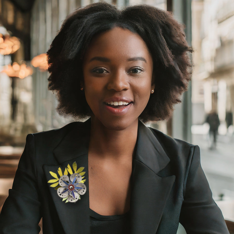 A stick pin featuring a silver flower and intricately cut iridescent gold leaves shown on a woman in a cafe wearing a black blazer