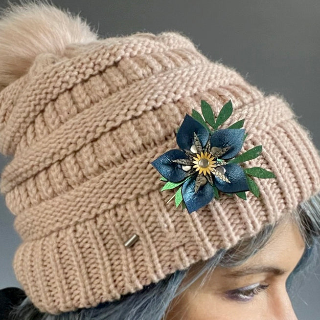 A stick pin featuring a blue flower and intricately cut iridescent green leaves shown on a pink knit hat