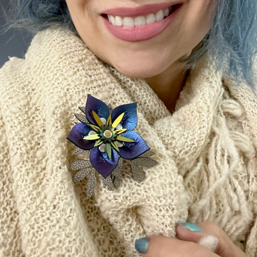 A stick pin featuring an iridescent blue flower and intricately cut gold flowers shown on a cream shawl