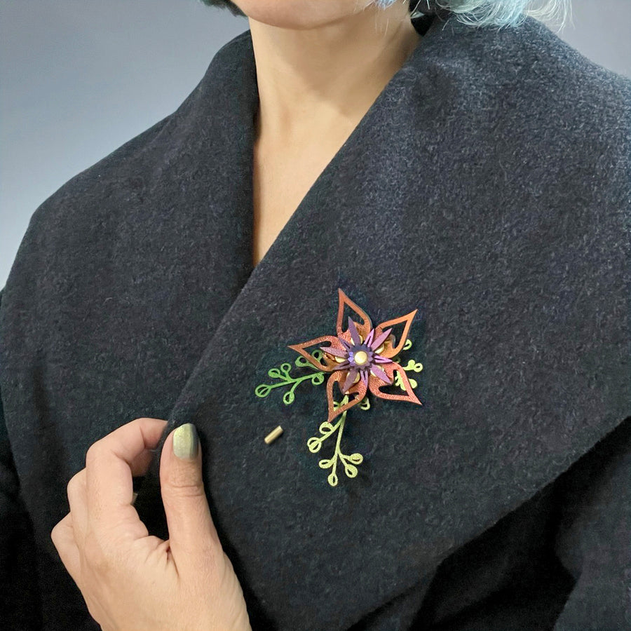 A stick pin featuring a red-bronze iridescent flower and intricately cut iridescent green leaves shown on a vintage charcoal coat