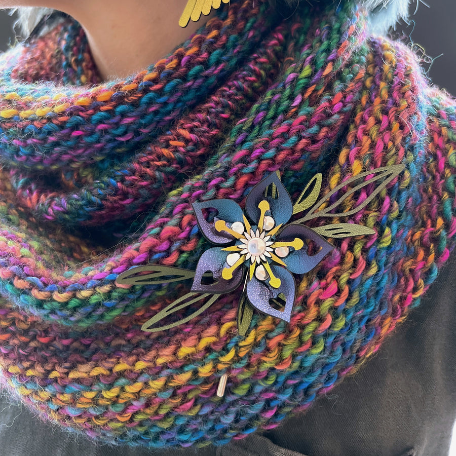 A stick pin (affixed to a colorful handmade scarf) featuring a 3D laser cut flower in shades of green, teal, purple , silver and gold