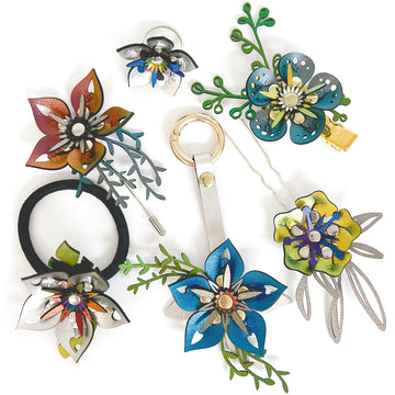 An assortment of 3D flower jewelry and accessories, including a ring, hair tie, barrette, stick pin, purse charm, and hair pin. Shown in a variety of iridescent vegan leathers